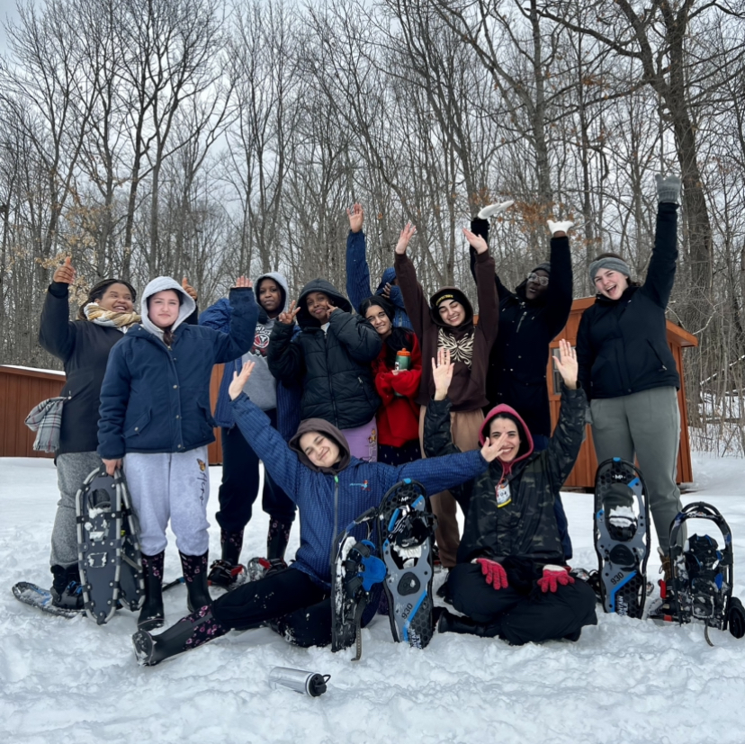 A group of youth are posing for a photo. They are all bundled up in winter clothes, there are snowshoes pitched in the snow around them. Some youth are sitting in the snow and some are standing behind them. They all have their arms up in the air.