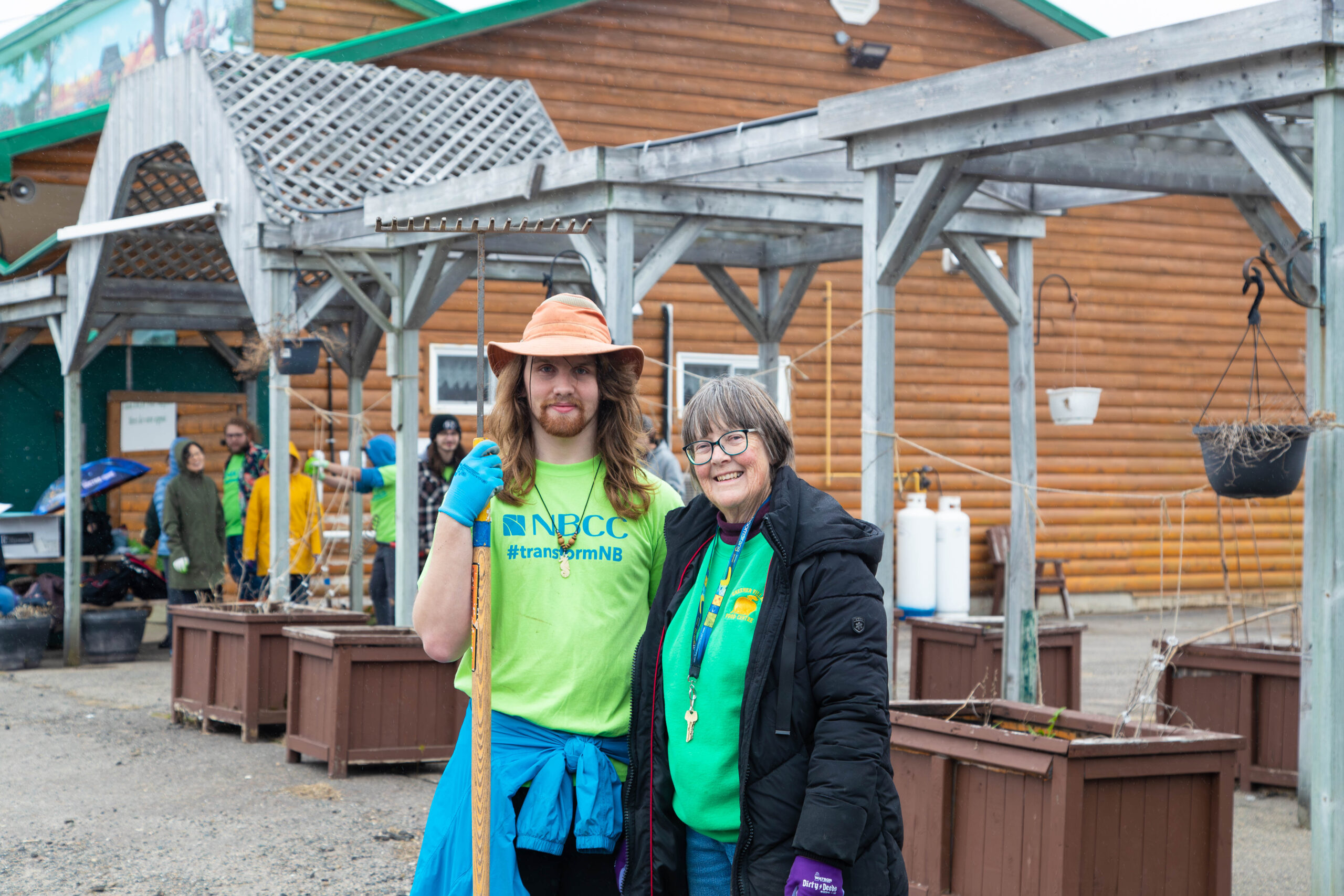 Two people stand together in front of a plant nursery. One person if holding a rake.