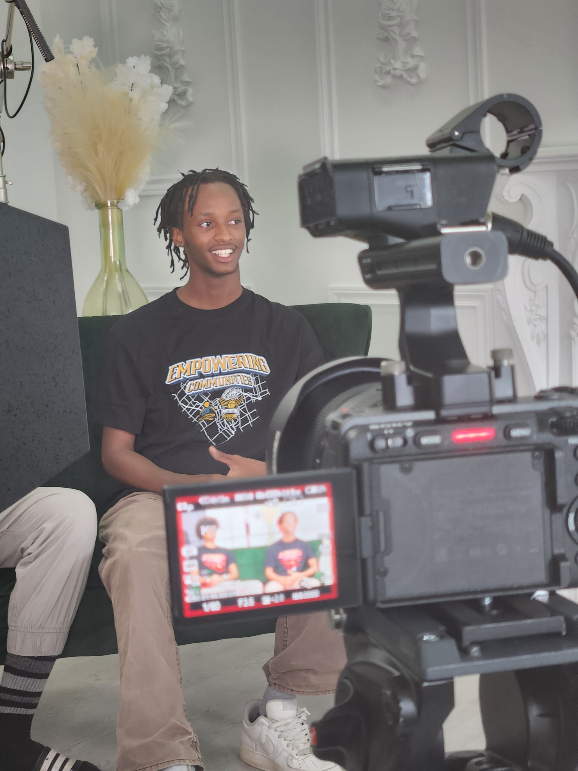 A young person smiles while sitting in front of a broadcasting camera.