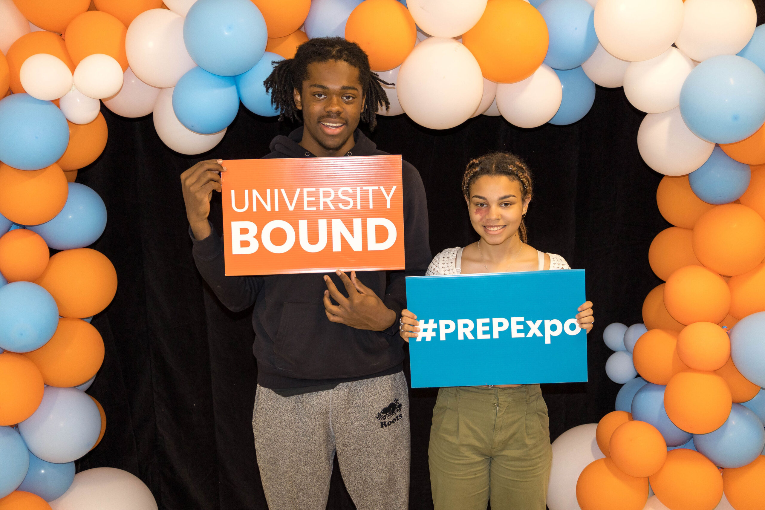 Two young people stand in front of a balloon arch. The balloons are white, orange, and blue. Both people are smiling and holding signs about PREP Academy.