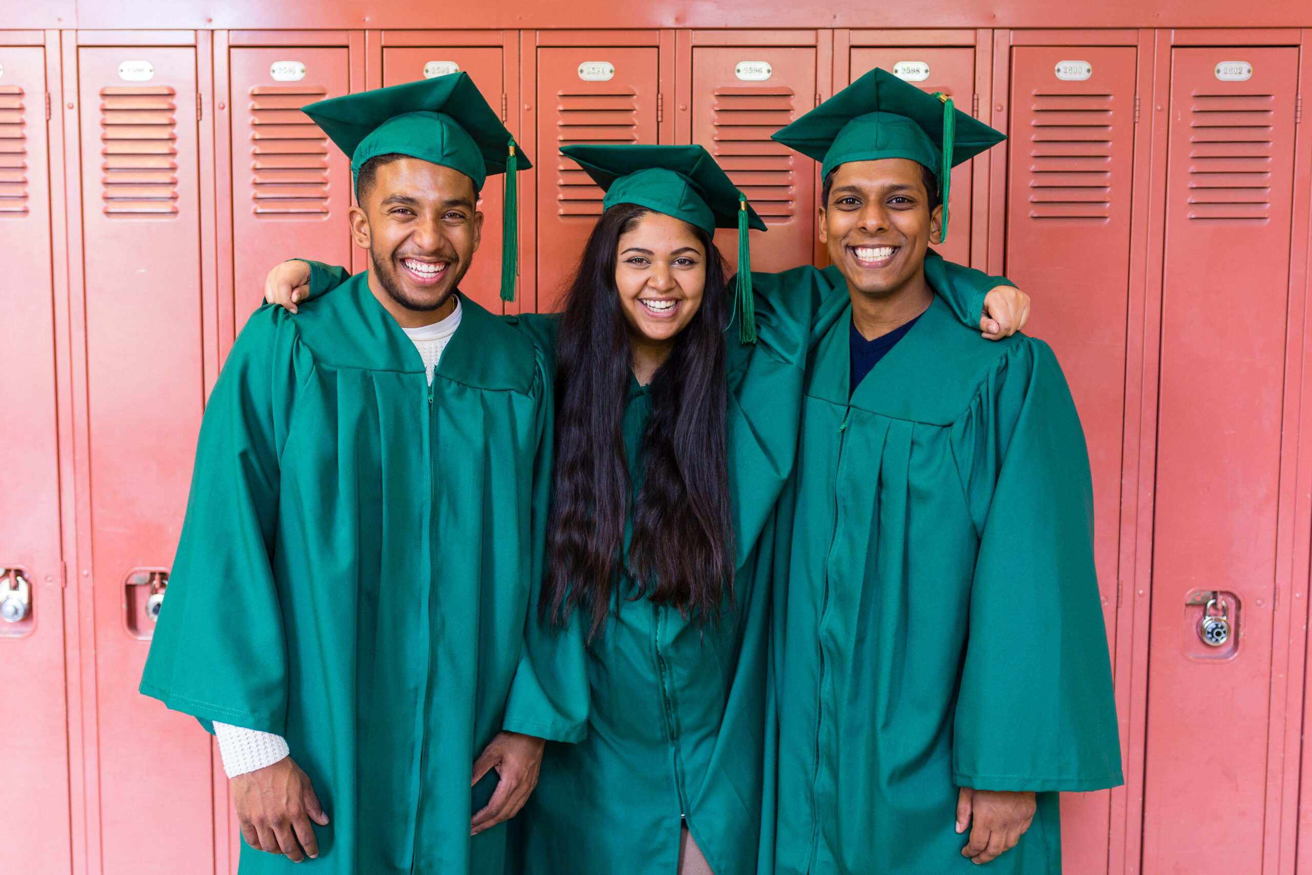 Three graduates stand together in front of orange school lockers. They are wearing dark green caps and gowns.
