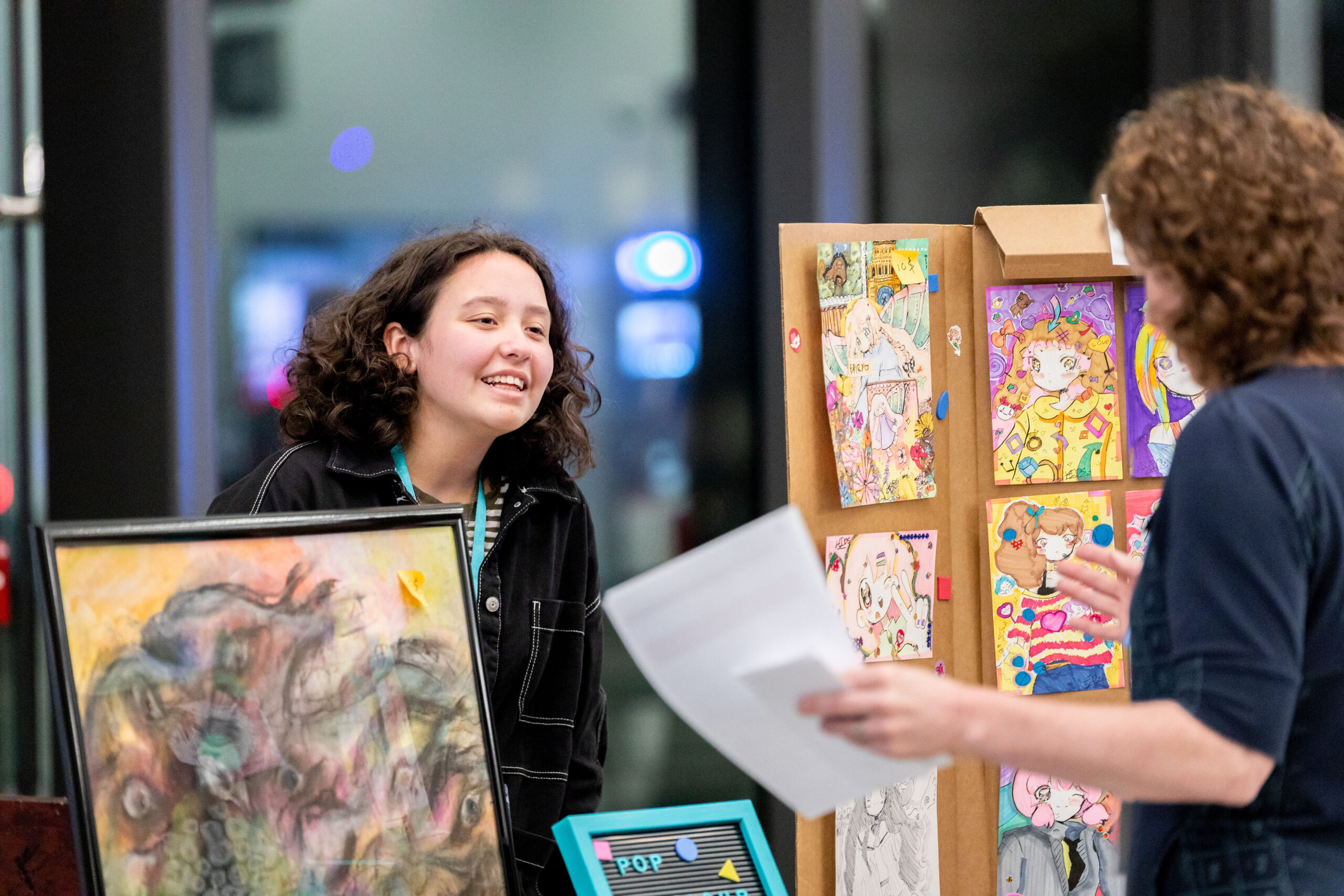 A young person stands behind a booth full of artwork. They are smiling at another person across the booth.